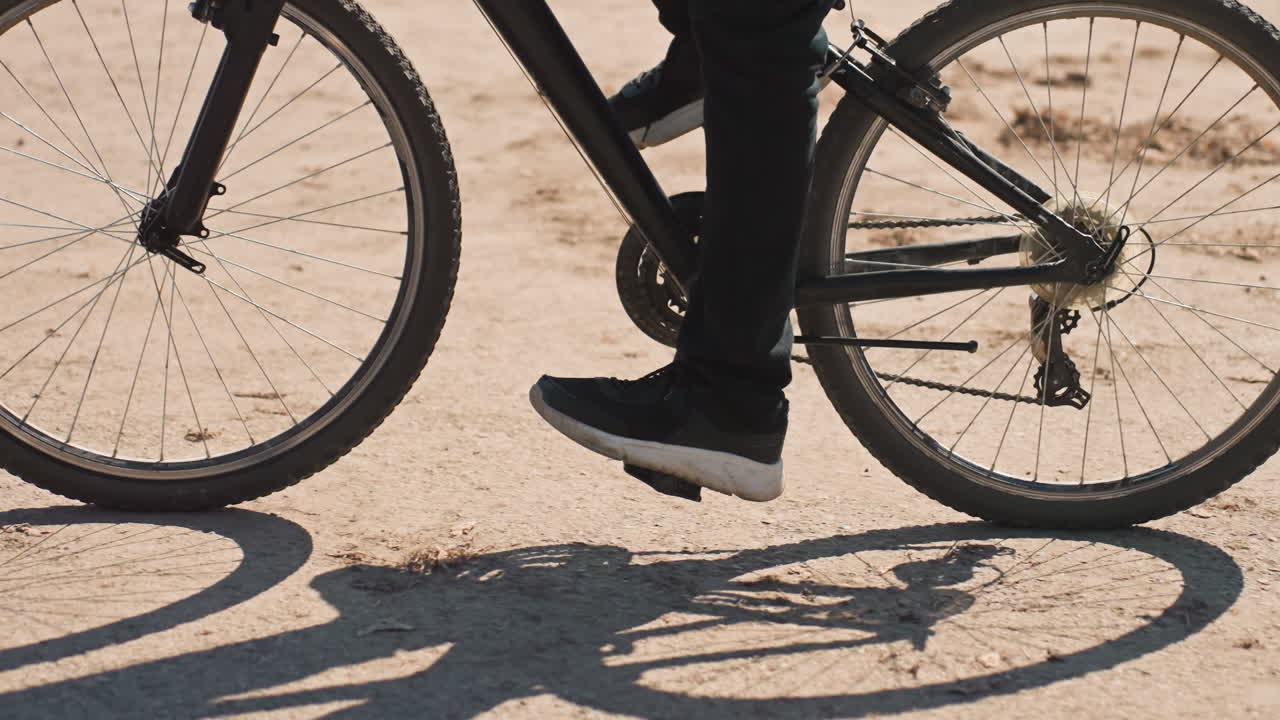 Cykelbud træder i pedalerne på støvet vej. En sort skygge af cykelhjul og sneakers er synlig. Grusbelægning, roterende kæde og eger fanger det varme sollys, hurtig levering, bylogistik, udendørs bevægelse