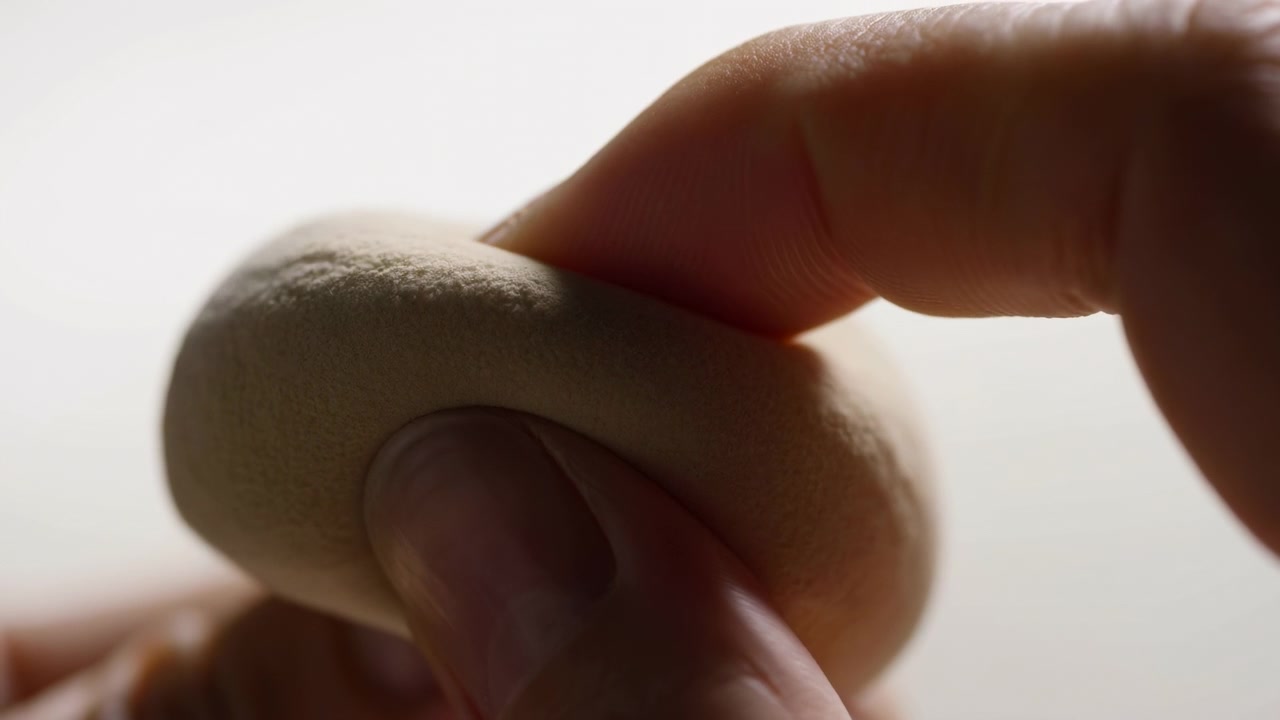 Close-up view shows fingertips squeezing a soft beige sponge while the porous material deforms and breaks apart, scattering tiny crumbs and powder through strong backlighting against pale space.