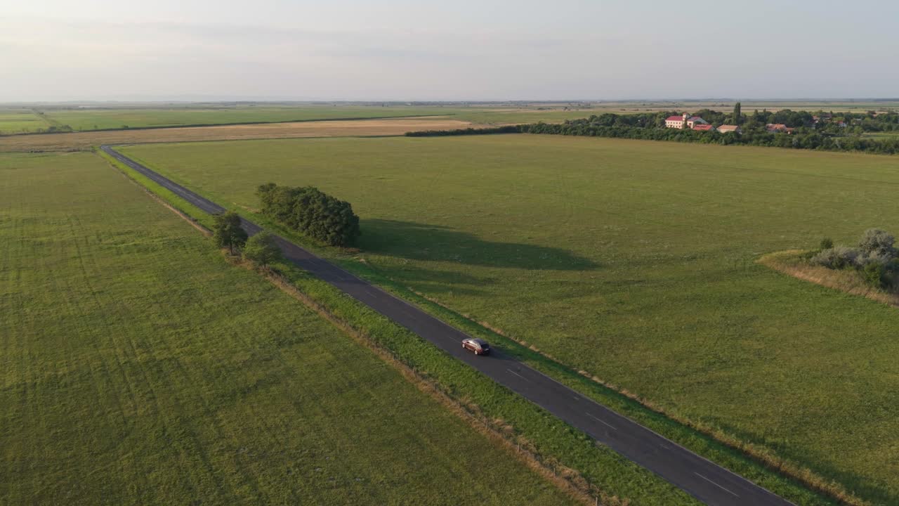 An aerial view of a single car traveling along a narrow rural road surrounded by vast green fields. The open landscape stretches to the horizon under a clear afternoon sky, creating a peaceful scene