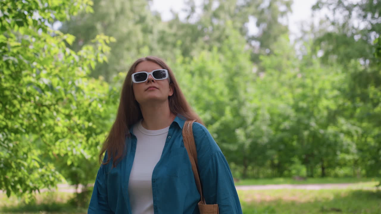 Female celebrity in stylish dark shades strolling along lush green park under bright daylight, wearing turquoise shirt and white top, exuding relaxed confidence and calm urban fashion