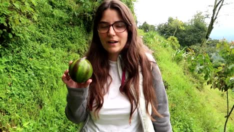 young woman with a fresh avocado in her hand talking about it during trekking in tropical green environment