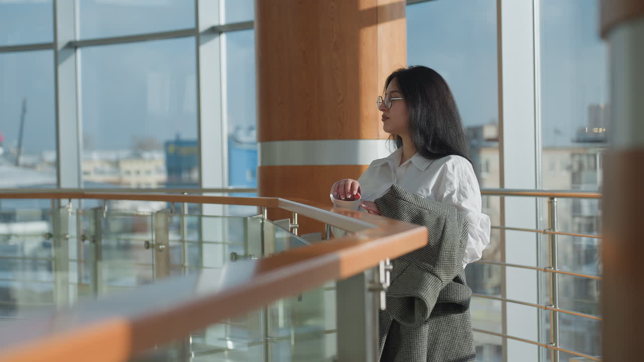 Confident female banker holding checkered coat walks along mall gallery, pauses to observe surroundings and rests on sleek railing with thoughtful gaze, framed by glass walls and wooden pillar
