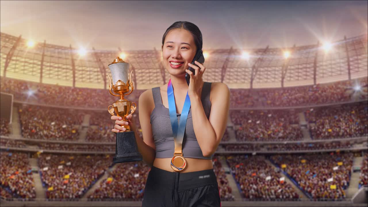 mujer asiática con una medalla de oro sosteniendo un trofeo de oro y hablando en un teléfono inteligente en el estadio