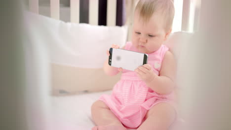 Little-baby-girl-with-phone-in-crib.-Infant-watching-cartoon-on-mobile-phone