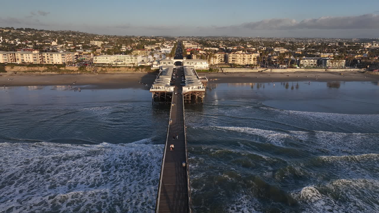 Drone shot flying straight down pier from ocean towards city beyond.