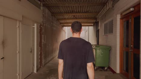 backview of a man slowly walking in the alley with green trash bin on the side - close up