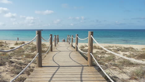 turista masculino con bermudas, polo, mochila y gorra caminando sobre un puente de madera procedente de una hermosa playa azul