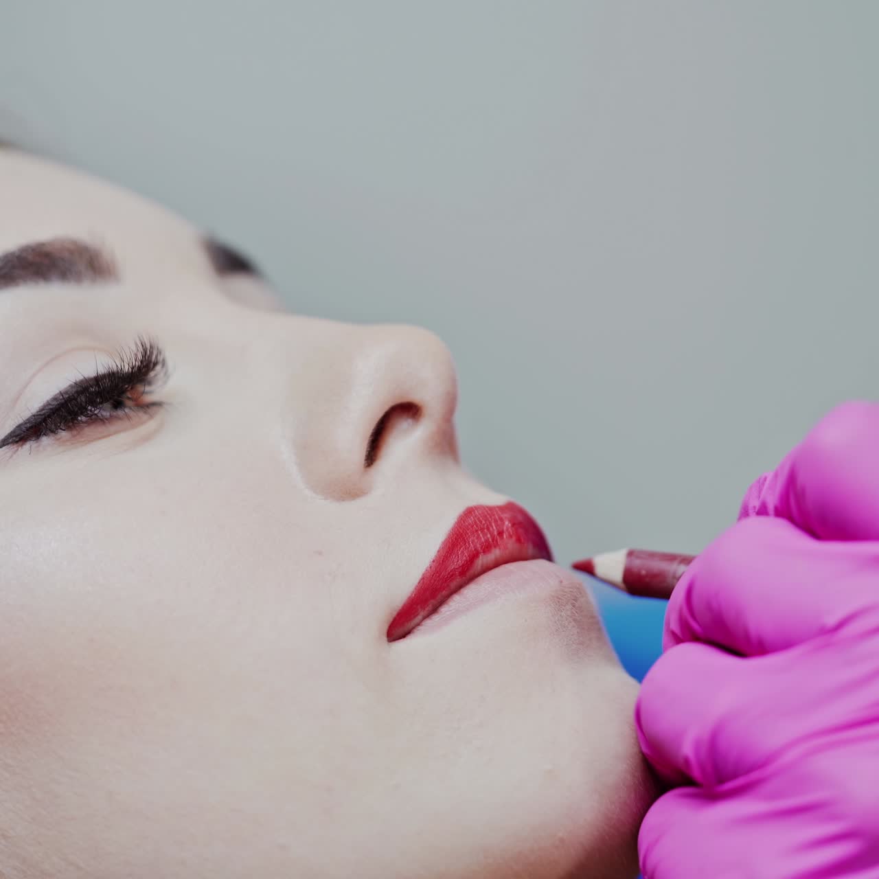 Permanent lip make-up in beauty salon. Cosmetologist draws the lips with a red pencil on the face of a young girl. Close-up.