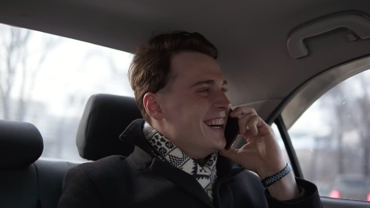 un chico atractivo y sexy con un abrigo elegante negro está sonriendo mientras habla con alguien por teléfono en un coche.