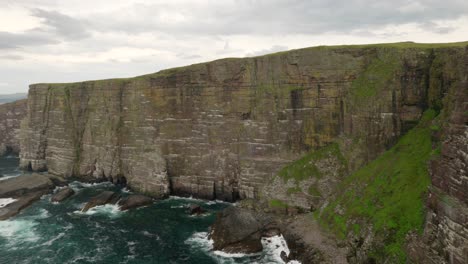 a slow panning shot revealing seabirds flying over a turquoise green ocean in front of a dramatic sea cliff rising straight up out of the ocean as waves crash against its base