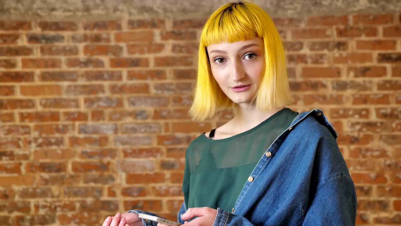 joven inusual con cabello amarillo sosteniendo el teléfono y mirando a la cámara, sonriendo y de pie cerca del fondo de la pared de ladrillo