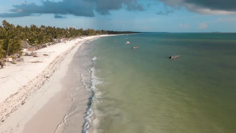 Aerial-view-of-sea-waves,-umbrellas,-palm-trees-and-walking-people-on-sandy-beach-at-sunset