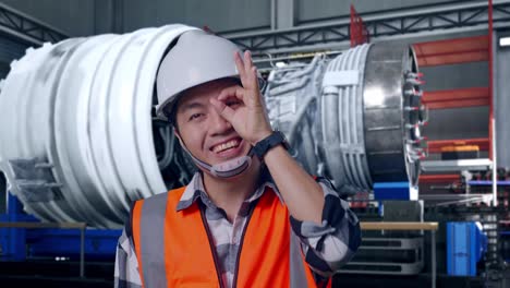 close up of asian male engineer with safety helmet showing ok hand sign over eye and smiling to camera while standing with airplane engine maintenance conducted, analytics checking the turbine