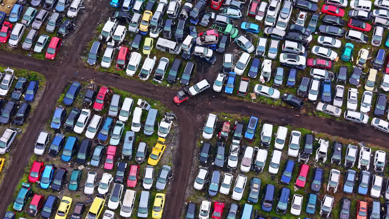 Diverse auto parcheggiate in un grande deposito di rottami situato fuori città durante le ore diurne, con sezioni visibili per diversi tipi di veicoli
