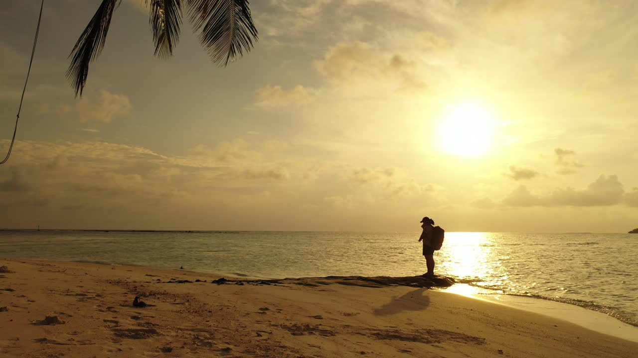 Backpacker standing over big rock on exotic beach under a palm tree at sunset moments with yellow sky reflecting on calm sea in Philippines