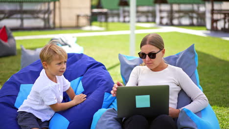 young mom and her little son sitting in bean bag chairs on the grass. woman shows something on laptop but the boy leaves.