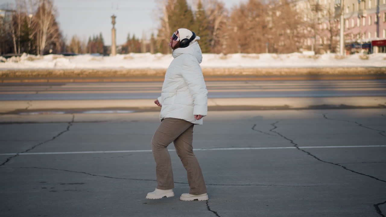 Side view of wandering performer in white winter coat dancing on cracked pavement, surrounded by snowy scenery and passing vehicles, expressing joy and freedom on cold urban street
