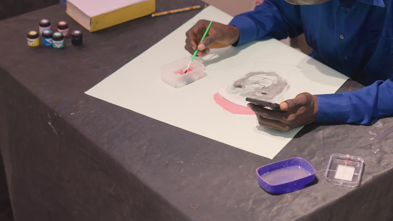 Black workshop instructor painting in studio, overhead workspace with jars, palette, phone reference and tools while artist applies black washes and pink accents, focused creative routine