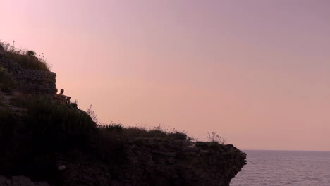 two silhouetted people sitting on cliff against backlit pink sunset sky in amalfi coast