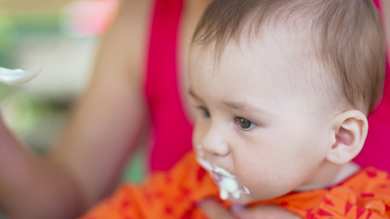 Little baby in orange sweater sits in mom's arms. Mother is feeding her kid from a spoon. Close up.