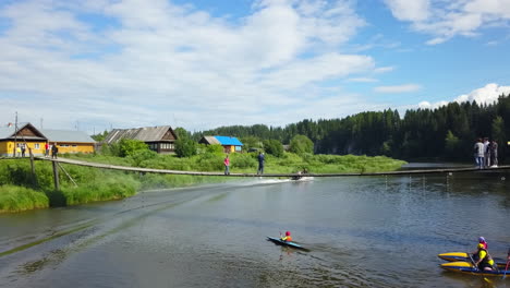 people enjoying kayaking and canoeing on a river with a bridge over it, in a picturesque village setting.