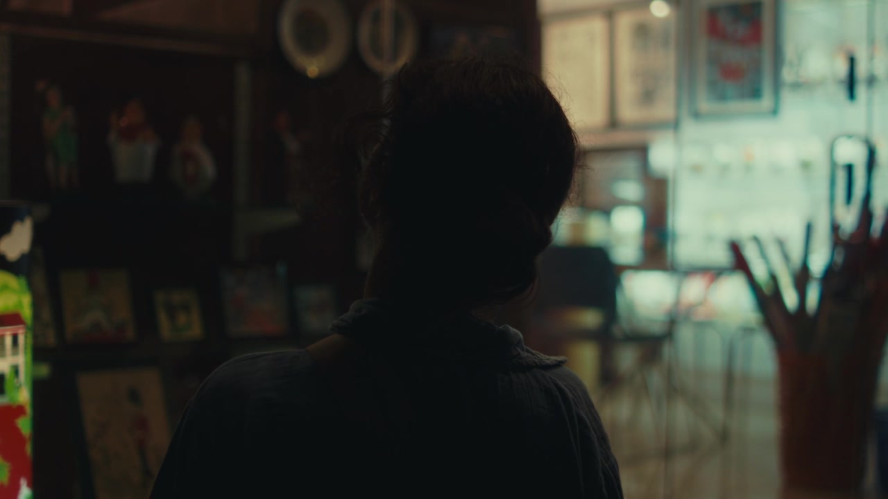 Woman exploring a dimly lit gallery or cultural interior, viewed from behind among artwork, displays, and warm ambient lighting in a cinematic travel and lifestyle scene.