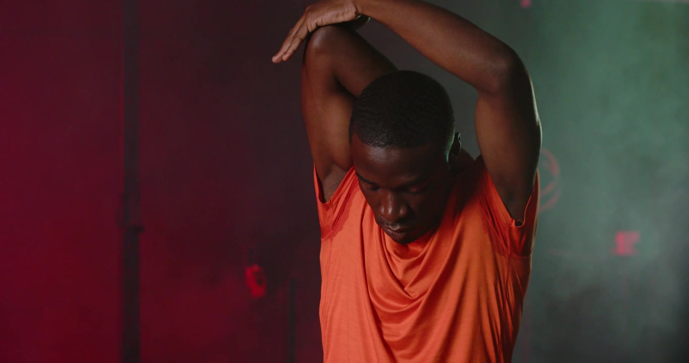 Man warming in studio in orange-shirt pulling right arm holding triceps stretch preparing workout