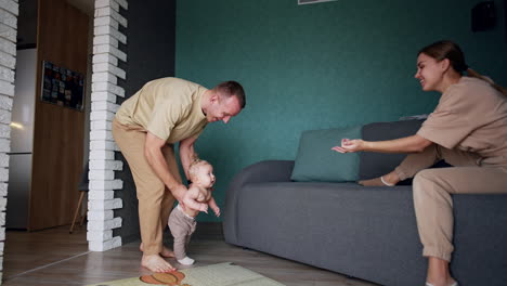 father holds a little baby trying to make first steps. mother sitting on the sofa calls her child to come.