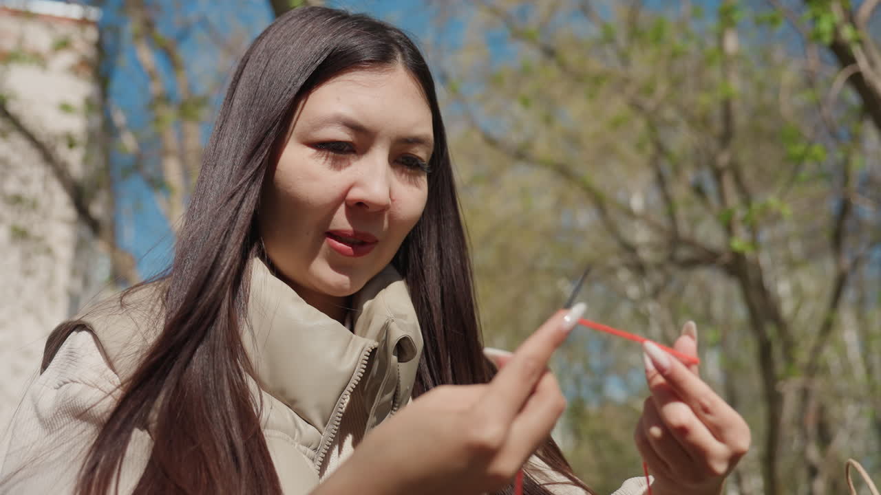공원에서 뜨개질하는 여성이 스마트폰 삼각대로 튜토리얼을 녹화하고 있어요. 빨간색 털실과 바늘로 작업하는 손을 클로즈업했으며, 햇빛이 나무 사이로 비치고, 집중한 표정으로 초보자를 위한 단계별 시연을 하고 있어요.