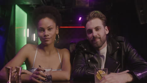 Multiethnic-Couple-Holding-Drinks-And-Smiling-At-Camera-While-Sitting-At-Bar-Counter