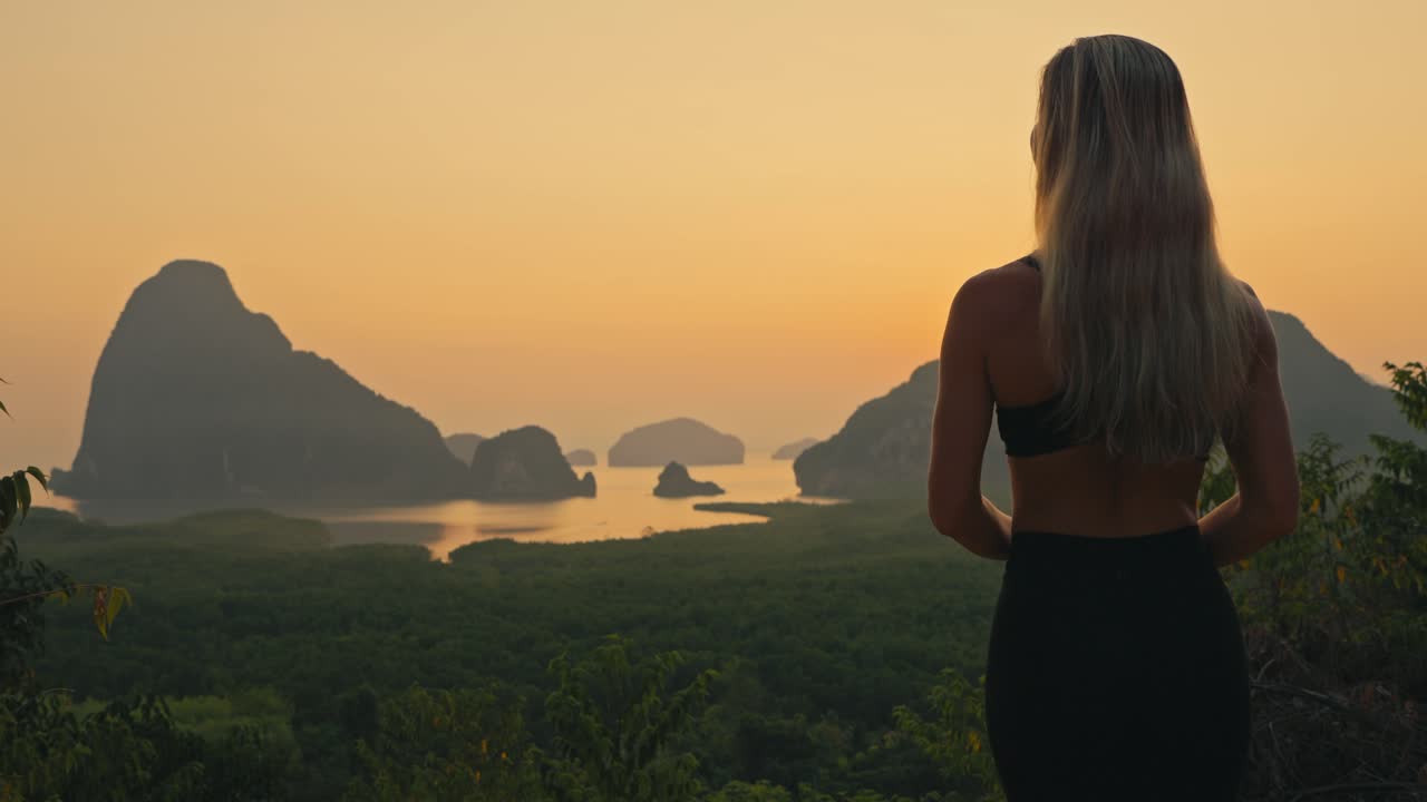 Beautiful fit blonde woman walks into frame and looks out over spectacular bay with limestone cliffs during sunrise with magical golden glow in the sky