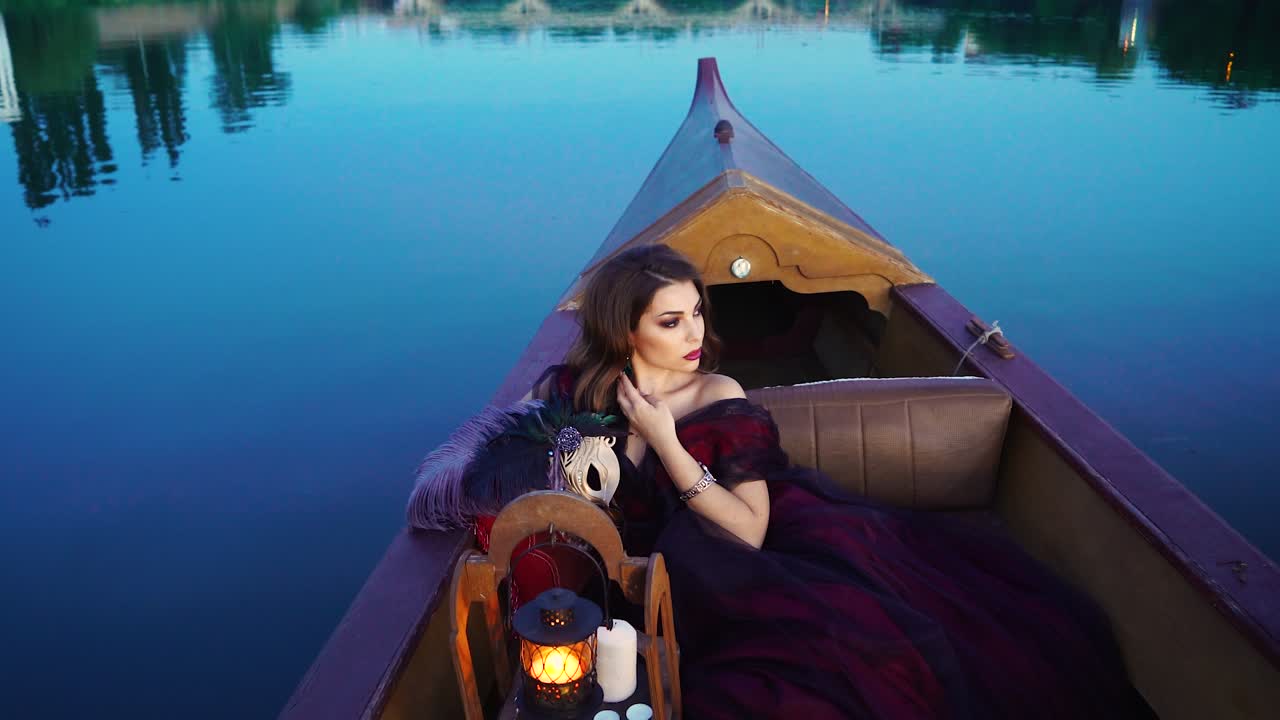 Gorgeous woman in long dark red dress sailing in a boat and touching her earing on blue water background. Beautiful model posing sitting in the gondola in the evening