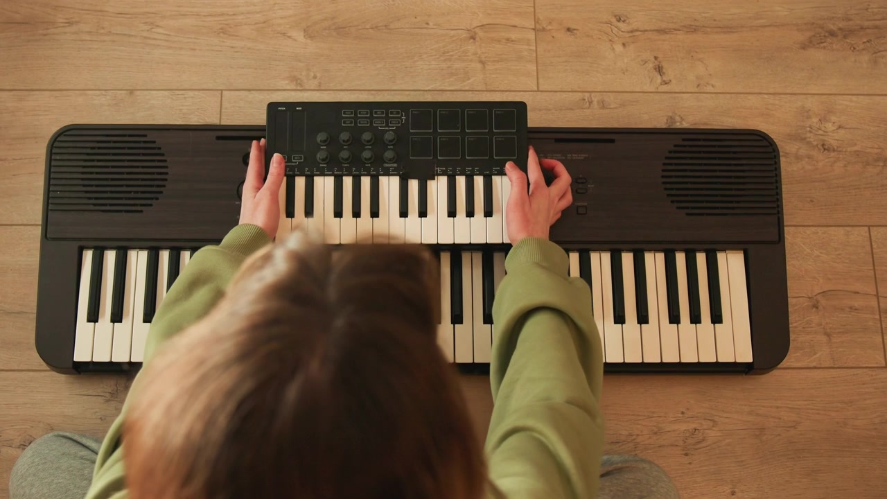 High overhead view of young child seated on hardwood floor playing compact electronic keyboard, with both hands pressing worn black and white keys and fingertips showing slight motion blur in warm