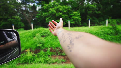 Caucasian-arm-hand-out-of-the-car-driving-fast-in-narrowed-road-with-green-vegetation-fresh-air-breeze-against-summer-heat