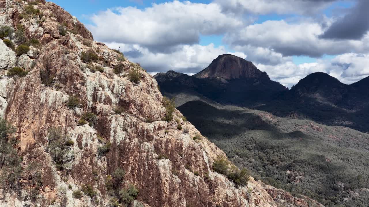 Camera smoothly pans along a rugged cliff face, revealing expansive mountain landscape and forested valley below, under bright daylight and dramatic clouds