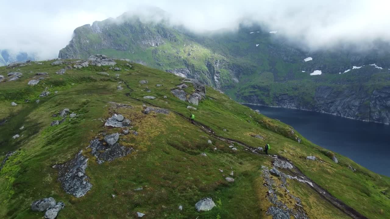 Aerial drone footage of two hikers with backpacks hiking at Lofoten Norway mountains and sheltered bays with a large blue lake in the background. Landscape is green, mossy and rocky and clouds are low