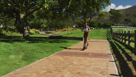 African-american-woman-wearing-headphones-and-backpack-riding-scooter-in-park