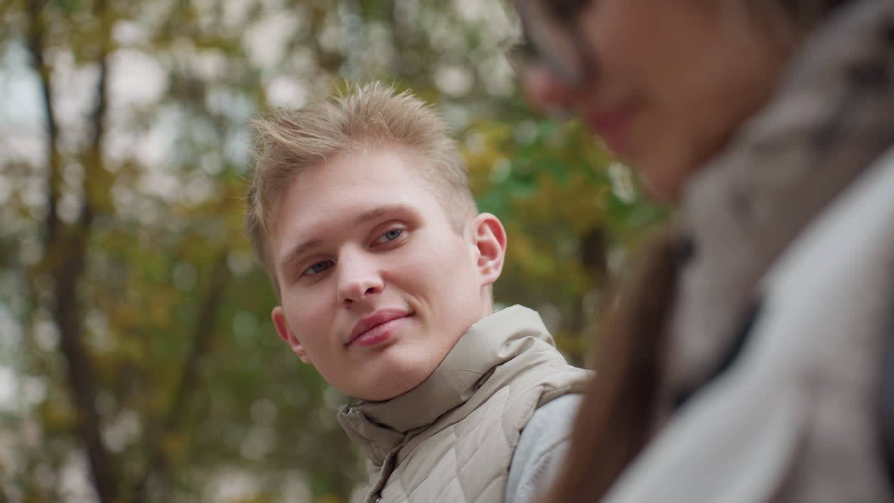 Close up of young lady wearing glasses with hair swaying in wind while walking outdoors, brother looking at her with soft expression, surrounded by blurred background of trees