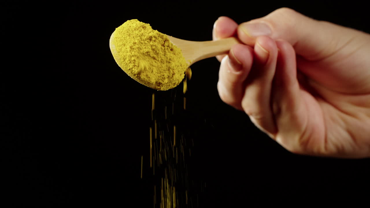 Hand Pouring Yellow Powder from Wooden Spoon on Black Background