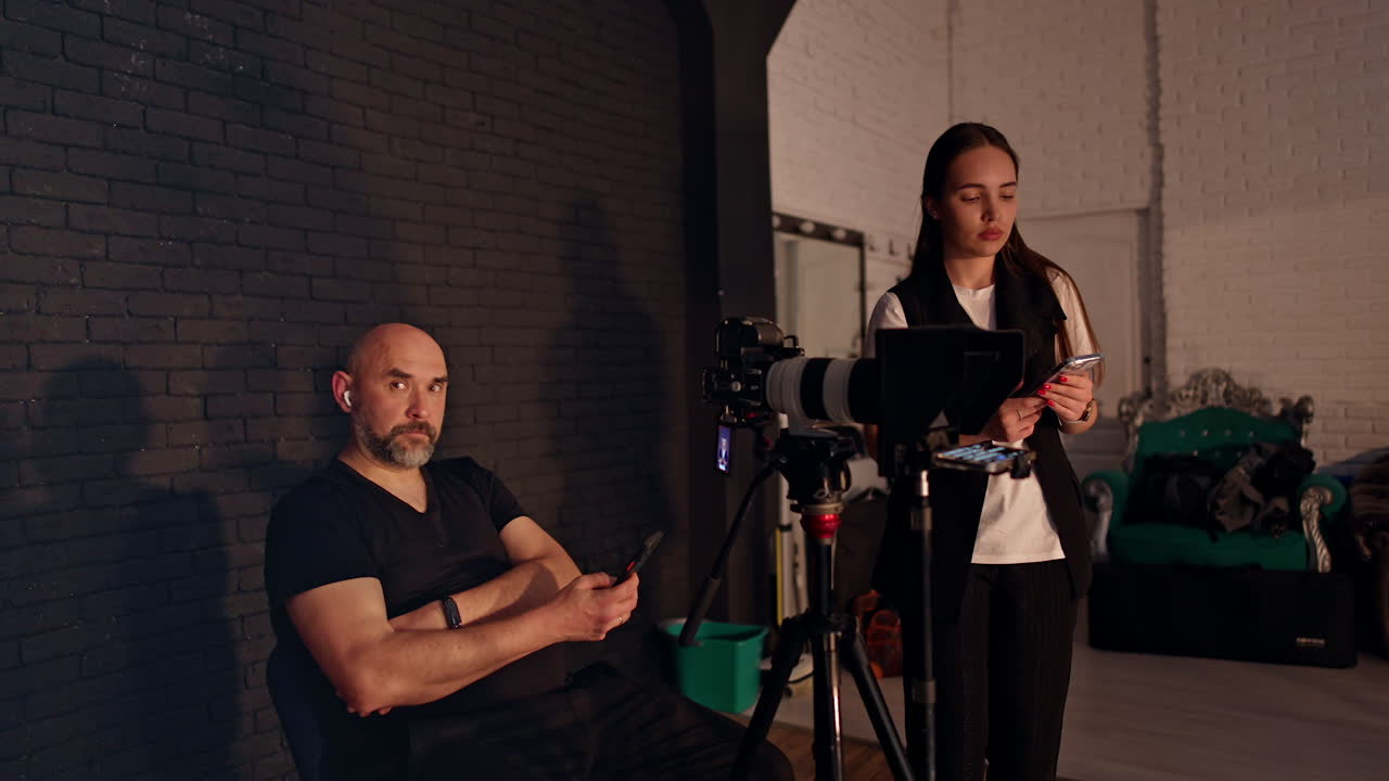 People assisting at blog content creation sit backstage in photo studio. Woman is taking video on her phone and man is looking at screen.