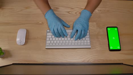 manos de una mujer con guantes azules protectores que escribe en el teclado en la oficina o en casa. teléfono inteligente con pantalla verde y tecla de croma con posición vertical de la pantalla. manos cercanas. cámara lenta