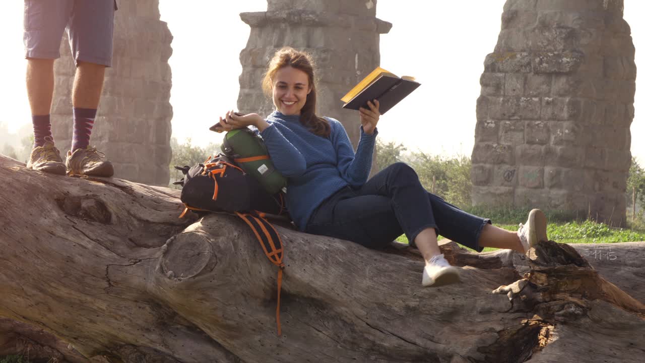 feliz mujer joven mochilero turistas sentados tendidos en un tronco de tronco leyendo un libro frente a las antiguas ruinas del acueducto romano en el parque degli aquedotti en roma al amanecer pareja romántica en cámara lenta