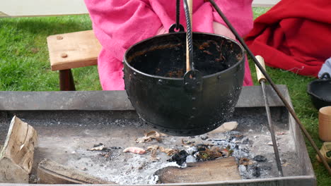 iron cauldron hanging over ash-filled fire pit, with remnants of a meal, capturing the essence of historic medieval viking outdoor cooking