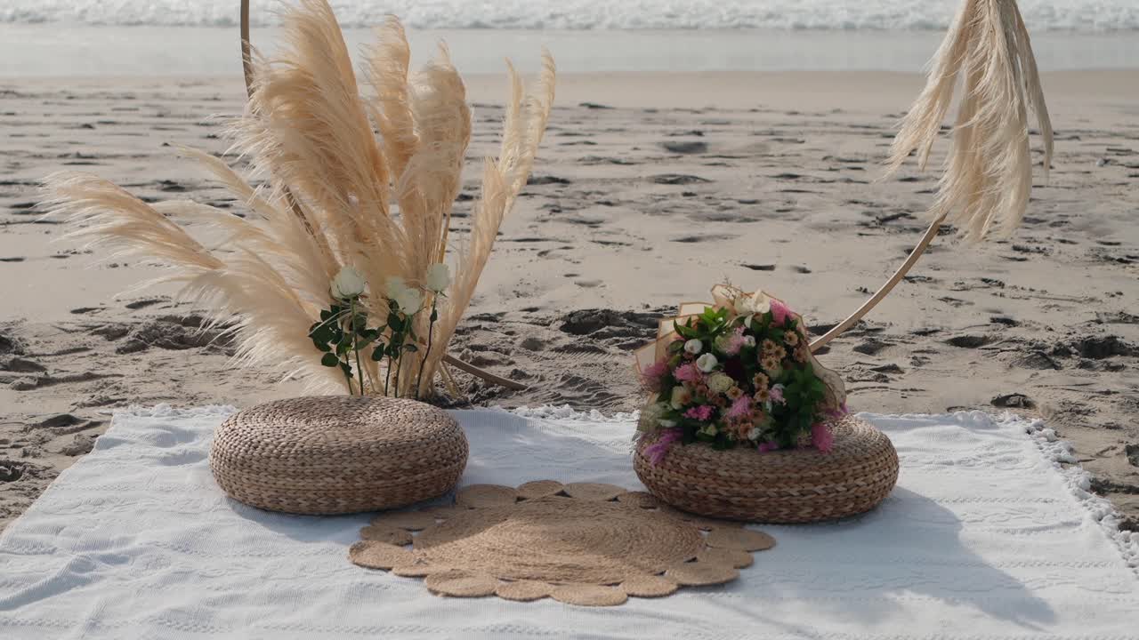 Boho wedding picnic decor on beach with straw pillows bouquet and pampas grass in soft light