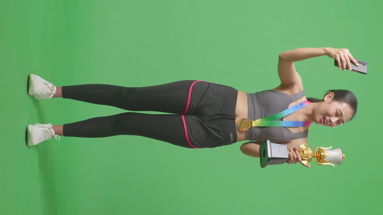 Full Body Of Asian Woman With A Gold Medal Showing A Gold Trophy To The Video Call On Smartphone On Green Screen Background In The Studio