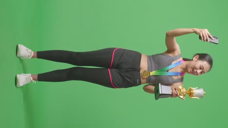 cuerpo lleno de mujer asiática con una medalla de oro que muestra un trofeo de oro a la llamada de video en el teléfono inteligente en el fondo de la pantalla verde en el estudio