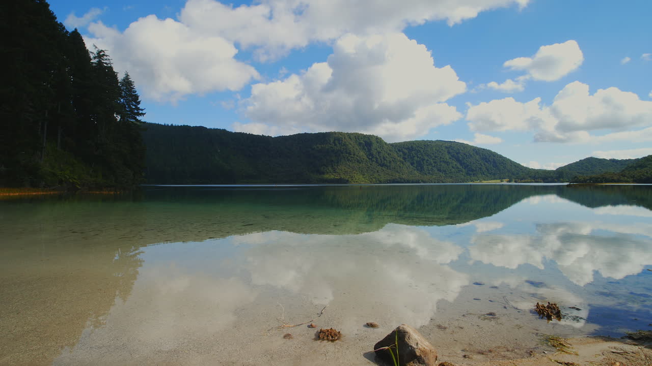 Pan left across reflecting Green Lake on a calm, partly cloudy day with blue skies in Rotorua, New Zealand