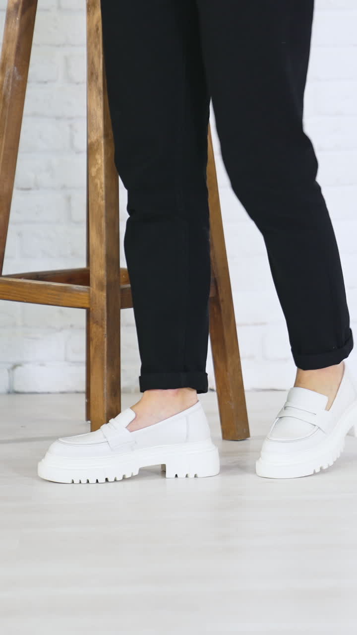 Girl in jeans and white fashionable sneakers waving her feet sitting on the step. Modern footwear demonstration in studio. Vertical video