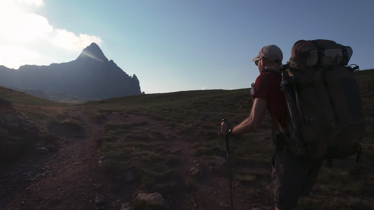 vista trasera de un excursionista con una gran mochila caminando hacia el pico de la montaña en la soleada puesta de sol de la tarde en verano