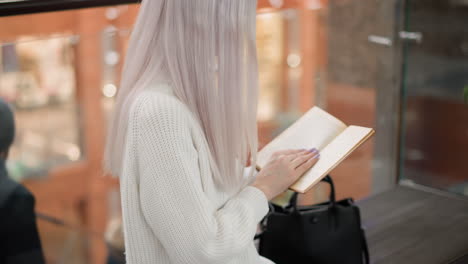 back view of contemporary woman in cozy sweater seated on mall bench turning pages of novel with care while people pass on moving walkway in background creating quiet peaceful reading moment